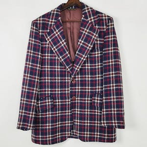 May include: A vintage blazer with a plaid pattern in navy blue, red, and white. The blazer has a notched collar, two front buttons, and two front pockets. The lining is a burgundy color. The blazer is hanging on a wooden hanger.
