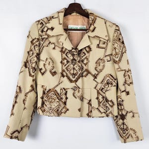 May include: A beige jacket with a brown geometric pattern. The jacket has a collared neckline, two front pockets, and a button closure. The jacket is hanging on a wooden hanger against a white background.