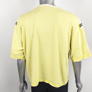 May include: A yellow short-sleeved t-shirt with black and white trim on the shoulders and sleeves. The t-shirt is on a mannequin.