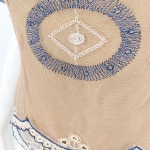 May include: A beige mesh top with a blue and white embroidered design. The top has a scalloped lace trim.