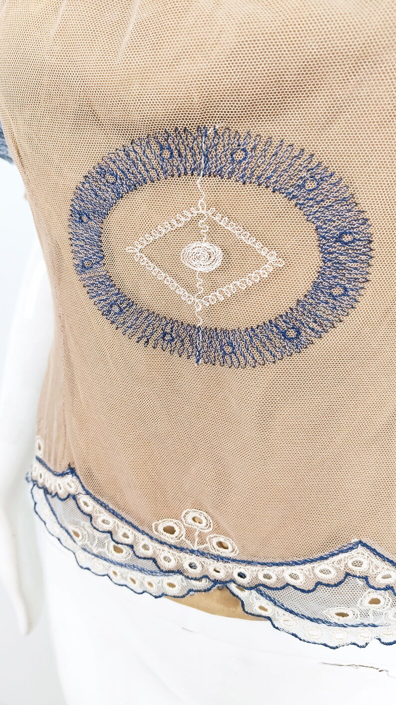 May include: A beige mesh top with a blue and white embroidered design. The top has a scalloped lace trim.