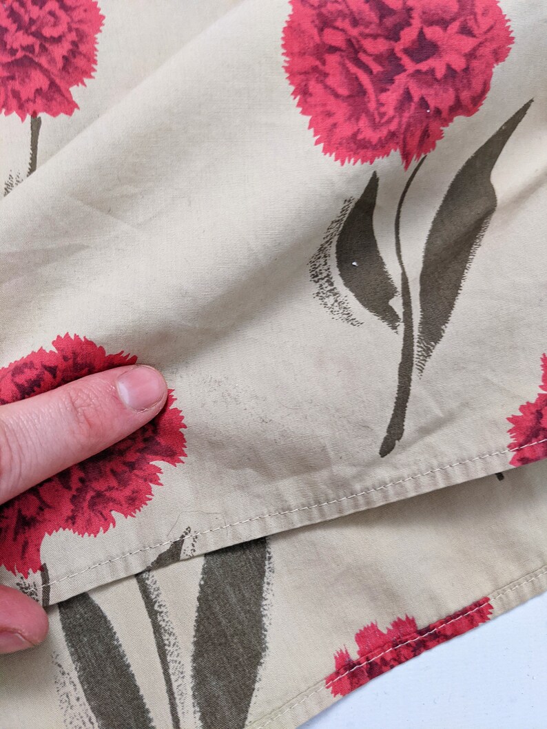 May include: Beige fabric with a repeating pattern of red carnations and dark green stems. The fabric is folded over and stitched along the edges.