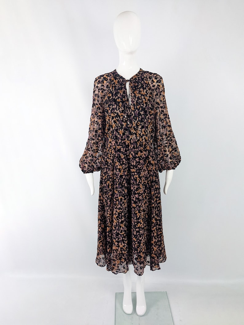May include: A long-sleeved midi dress with a black and brown abstract print. The dress has a gathered neckline with a tie detail and a button closure. The sleeves are gathered at the shoulders and the skirt is flowy.