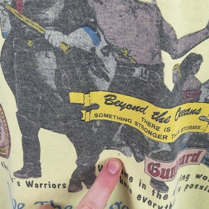 May include: A yellow t-shirt featuring a graphic print of two men holding weapons. The text on the shirt reads "Beyond the Jeans, There is something stronger than storms... Guard Warriors, Welcome here in the... Be the Legend!"