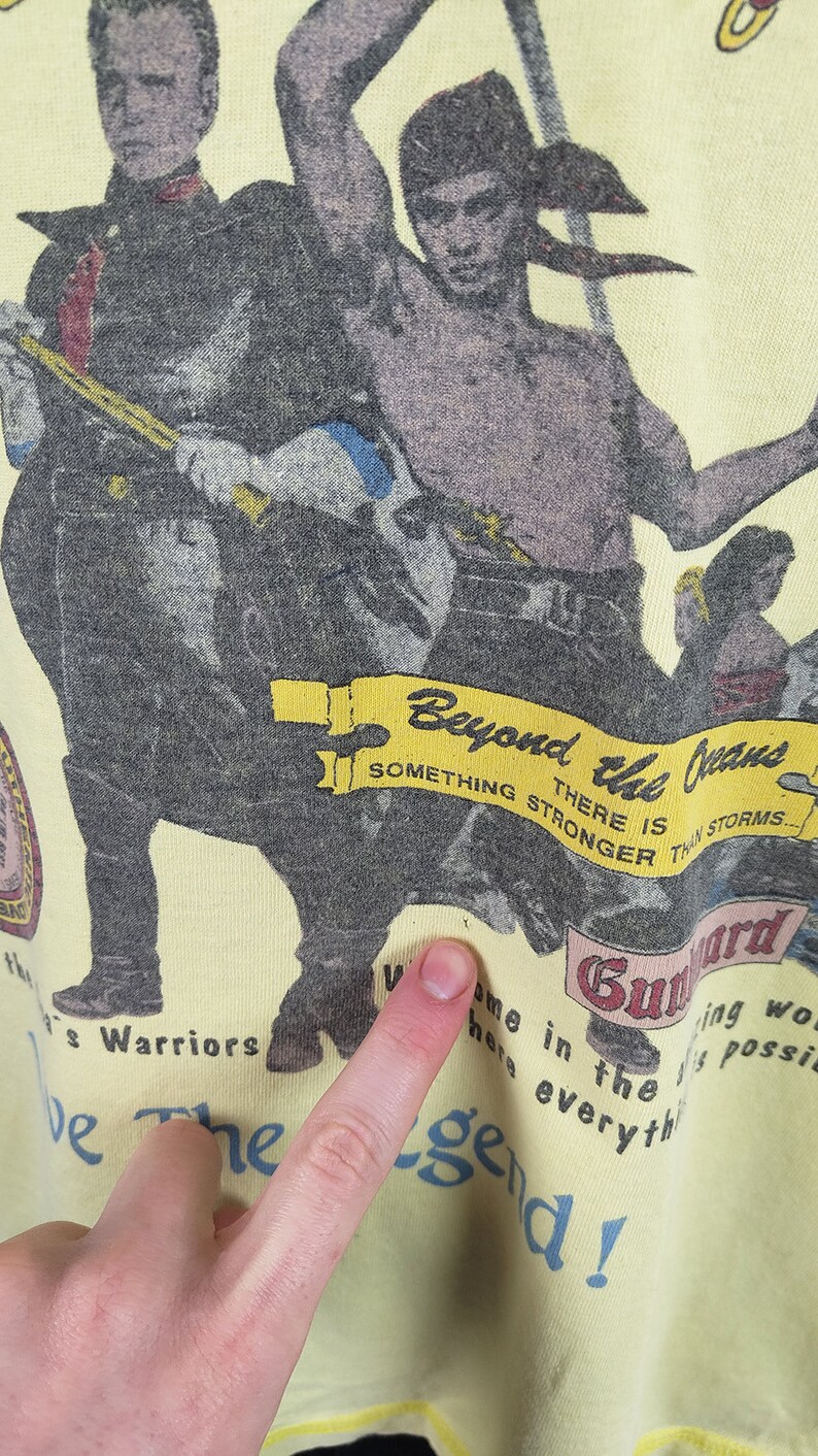 May include: A yellow t-shirt featuring a graphic print of two men holding weapons. The text on the shirt reads "Beyond the Jeans, There is something stronger than storms... Guard Warriors, Welcome here in the... Be the Legend!"