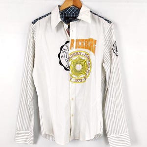 Puede incluir: Camisa blanca abotonada con mangas a rayas finas y cuello a cuadros. La camisa presenta un gran gráfico en la parte delantera con el texto "R ICEBERG ACADEMY - ICE PL 1973". Parches adicionales en las mangas.