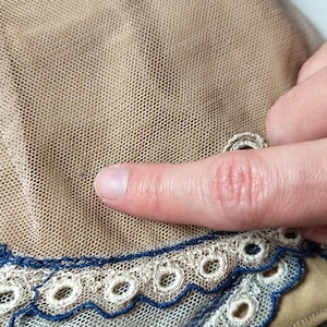 May include: Close-up of a beige mesh fabric with a white lace trim and a blue embroidered design. The fabric is soft and delicate, and the lace is intricate and beautiful.