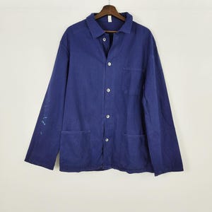 Puede incluir: Chaqueta de trabajo azul marino vintage con botones, dos bolsillos delanteros y un bolsillo en el pecho. La chaqueta está colgada de una percha de madera. Hay marcas de pintura en la manga izquierda.