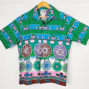 May include: A short-sleeved button-up shirt with a vibrant floral and geometric pattern. The shirt features a green collar and sleeves, with blue and pink accents. The design includes circular and floral motifs in shades of brown, purple, and white.
