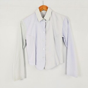 May include: A long-sleeve button-down shirt featuring a colour-blocked design with a light lavender centre panel and contrasting light grey sleeves. The shirt has a structured collar, a front button placket with white buttons, and a textured fabric.