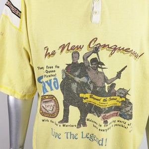 May include: Yellow t-shirt with a graphic print of pirates and the text "The New Conquery!"  They free the Queen Pirates! Furyo. Beyond the Oceans. Something Stronger Than Storms. Gunboard. Welcome in the racing world of here everything is possible! Live The Legend!"