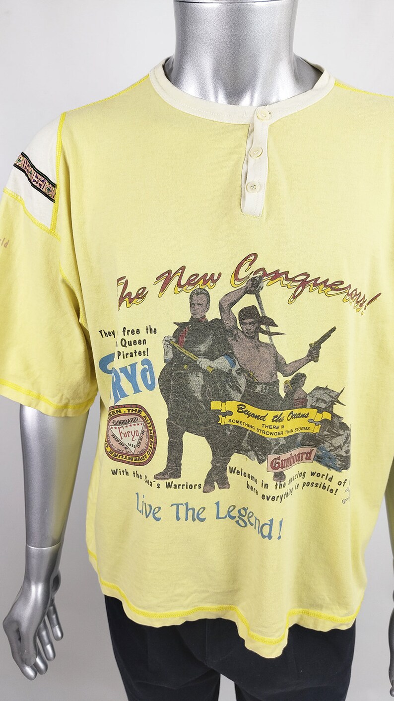 May include: Yellow t-shirt with a graphic print of pirates and the text "The New Conquery!"  They free the Queen Pirates! Furyo. Beyond the Oceans. Something Stronger Than Storms. Gunboard. Welcome in the racing world of here everything is possible! Live The Legend!"