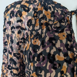 May include: A black, brown, and purple sheer blouse with a leopard print pattern. The blouse has a tie at the neck and long sleeves.