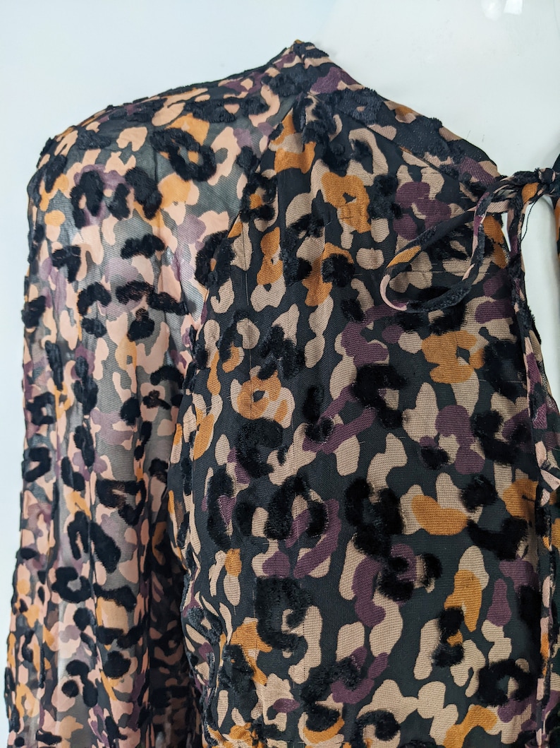 May include: A black, brown, and purple sheer blouse with a leopard print pattern. The blouse has a tie at the neck and long sleeves.