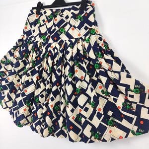 May include: A vintage skirt with a geometric pattern. The skirt features a cream-colored background with navy blue and white geometric shapes, accented by small orange squares and green floral designs. The skirt is pleated and hangs from a black hanger.