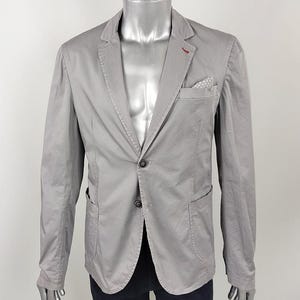 Puede incluir: Una chaqueta gris claro con un cierre de botón único y dos bolsillos. La chaqueta tiene un detalle de costura roja en el cuello.