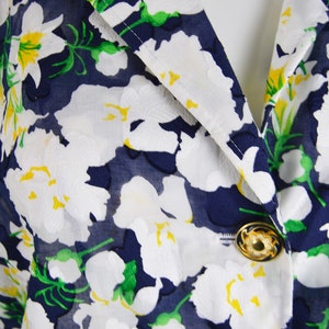 Pu&ograve; includere: Primo piano di un tessuto blu navy con un motivo floreale bianco. Il tessuto ha una trama trasparente e presenta accenti gialli nei fiori. Due bottoni dorati sono visibili sul tessuto.