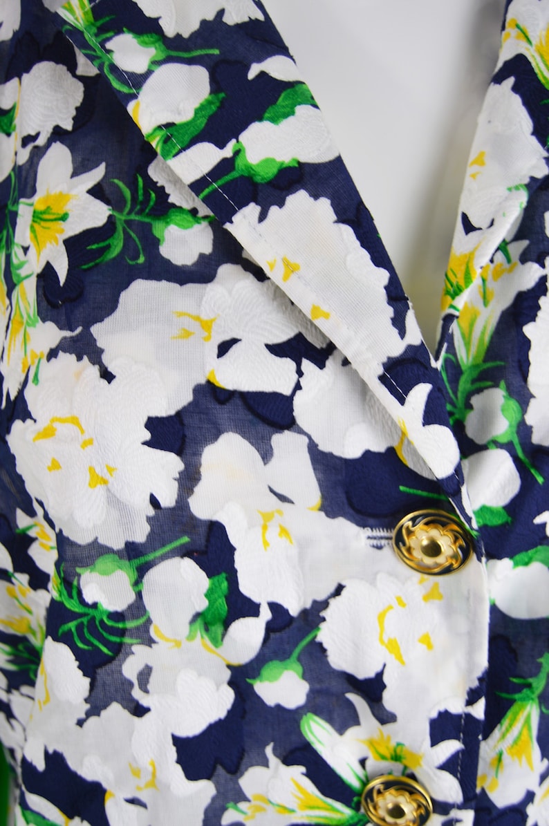 Pu&ograve; includere: Primo piano di un tessuto blu navy con un motivo floreale bianco. Il tessuto ha una trama trasparente e presenta accenti gialli nei fiori. Due bottoni dorati sono visibili sul tessuto.