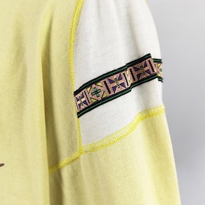 May include: A yellow sweatshirt with a white and yellow patterned trim on the sleeve. The trim has a geometric design with green, purple, and brown accents.