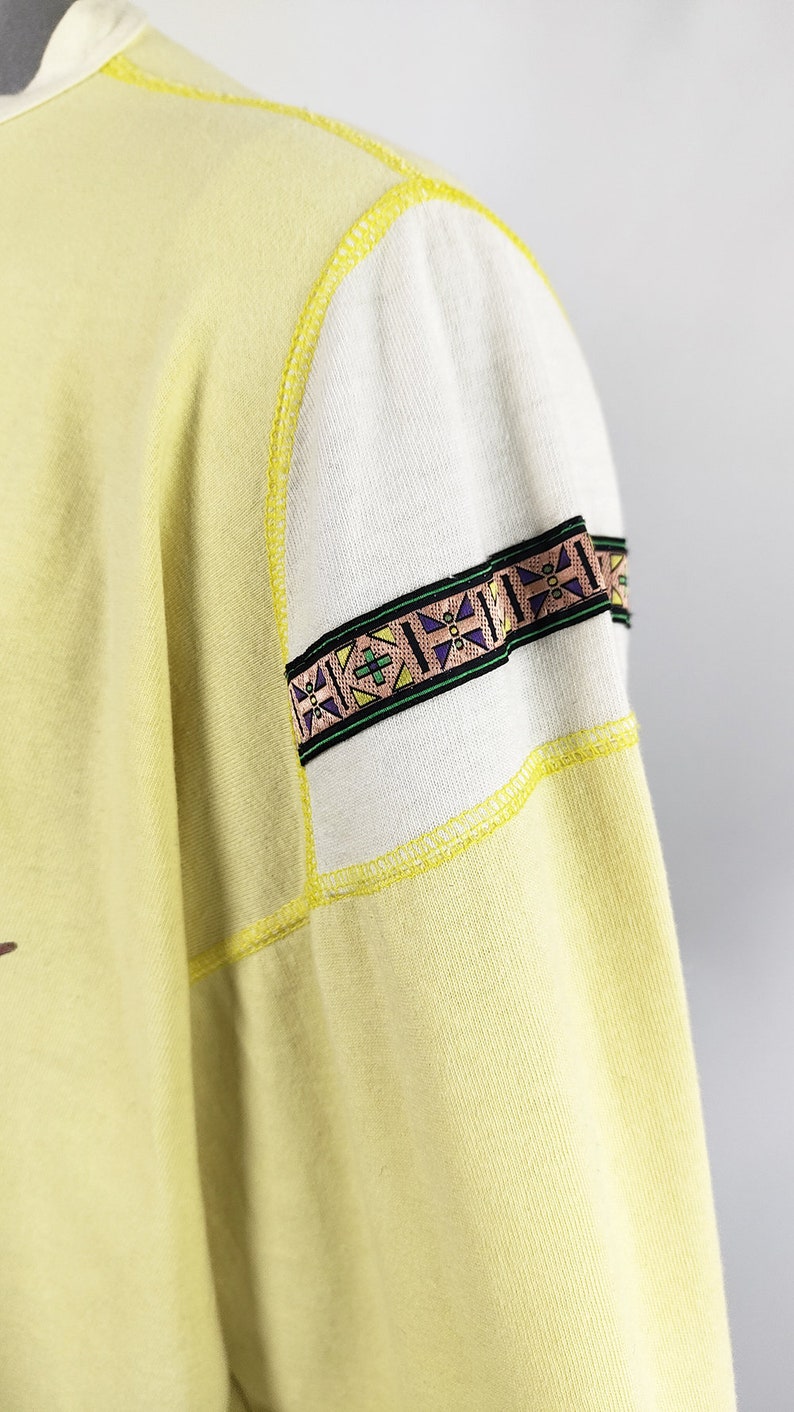 May include: A yellow sweatshirt with a white and yellow patterned trim on the sleeve. The trim has a geometric design with green, purple, and brown accents.