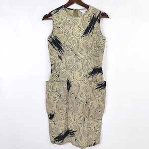 May include: Sleeveless dress with a beige paisley pattern and black brushstroke accents. The dress has a fitted silhouette, two front pockets, and a zipper closure. It is hanging on a wooden hanger against a white background.