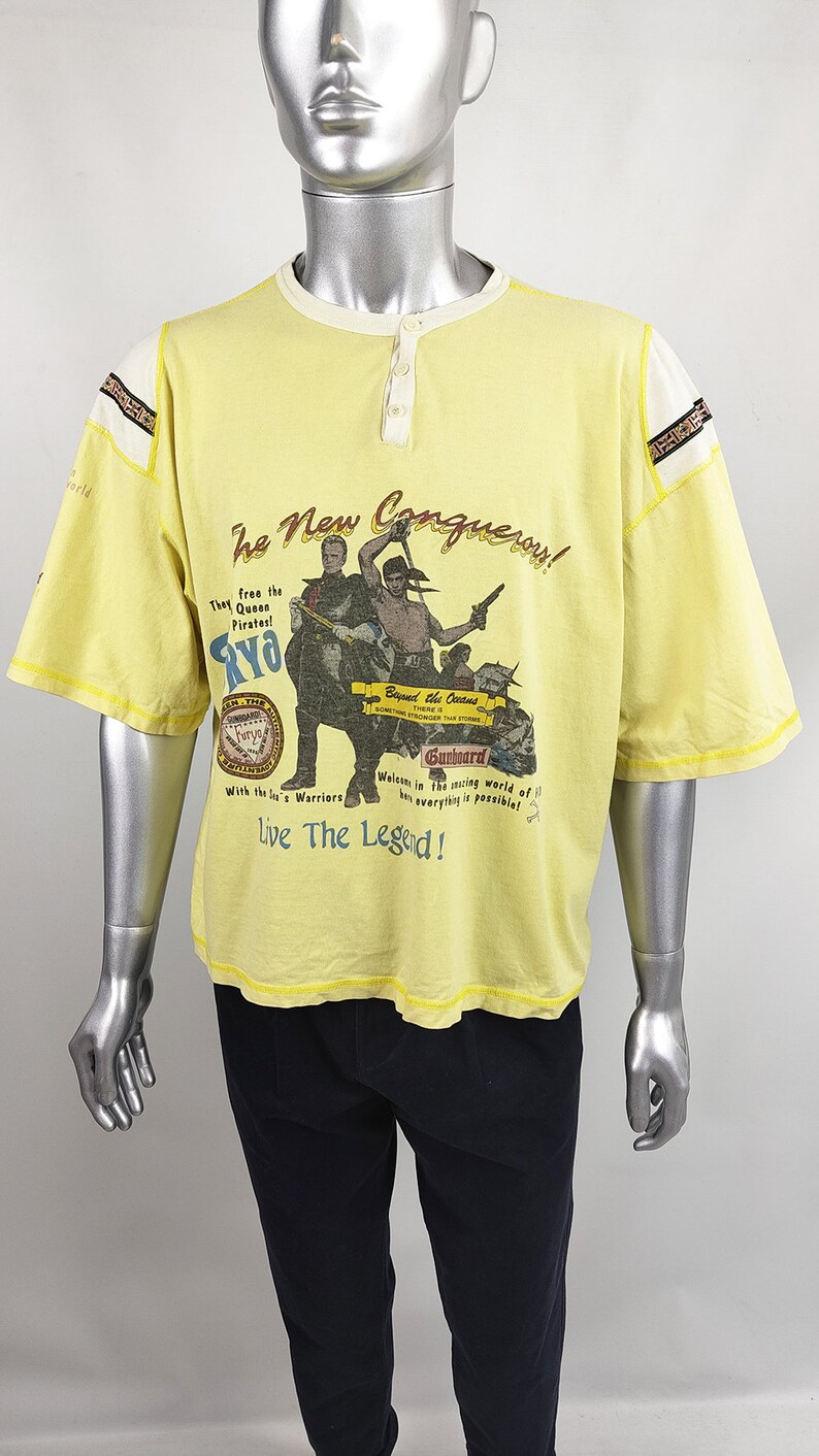 May include: Yellow t-shirt with a graphic print of pirates and the text "The New Conquerous!"  "They free the Queen Pirates! RYO"  "Beyond the Oceans There is Something Stranger Than Storms"  "Gunboard"  "Welcome in the exciting world of fun everything is possible!"  "Live The Legend!"