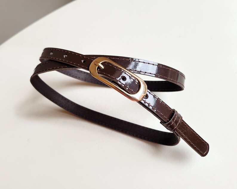 May include: A brown leather belt with a gold buckle. The belt is wrapped around itself and the buckle is visible in the centre of the image.