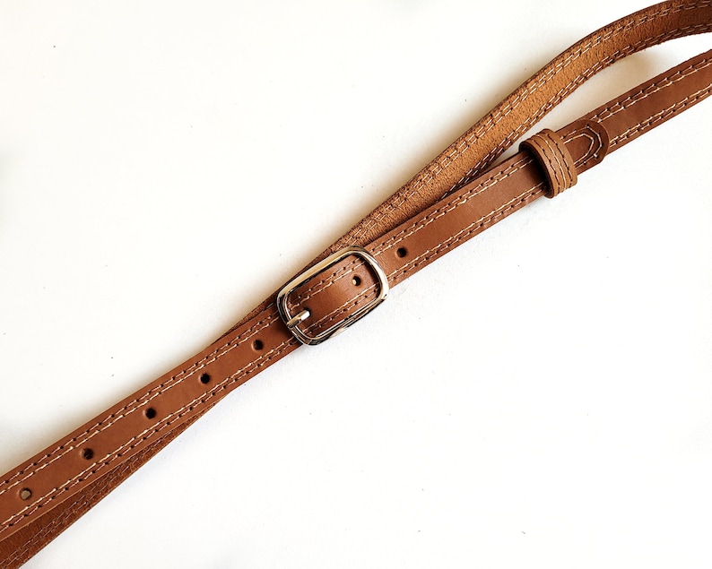 May include: A brown leather strap with a silver buckle. The strap is stitched with white thread.