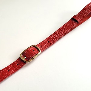 Puede incluir: Una correa de cuero roja con una hebilla dorada. La correa tiene un patr&oacute;n de piel de cocodrilo.