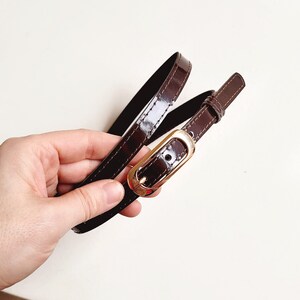 May include: A brown leather belt with a gold buckle. The belt is folded in half and held in a hand.