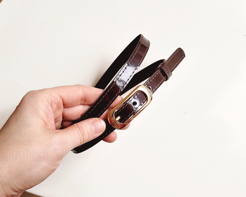 May include: A brown leather belt with a gold buckle. The belt is folded in half and held in a hand.