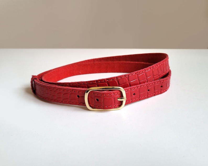 Puede incluir: Un cintur&oacute;n de cuero rojo con una hebilla dorada. El cintur&oacute;n est&aacute; estampado con un patr&oacute;n de cocodrilo.
