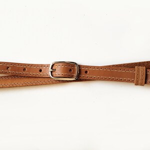 May include: A brown leather belt with a silver buckle. The belt is stitched with a white thread.