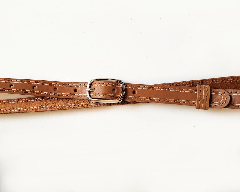 May include: A brown leather belt with a silver buckle. The belt is stitched with a white thread.