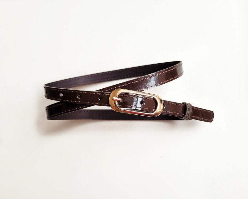 May include: A brown leather belt with a gold-toned buckle. The belt is folded in half and the buckle is visible in the centre of the image.