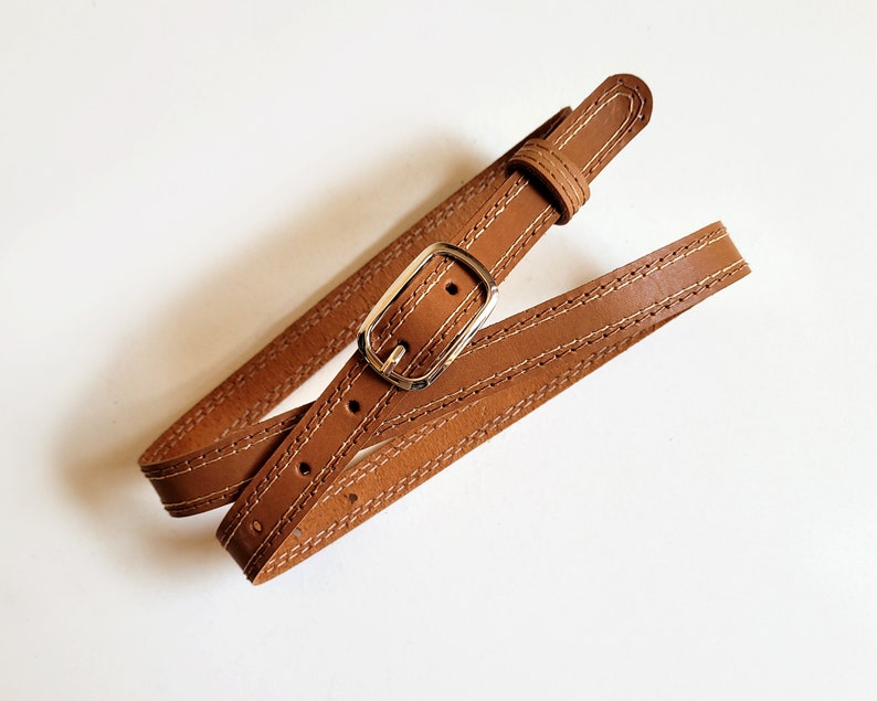 May include: A brown leather belt with a gold buckle and stitching. The belt is folded in half and the buckle is visible.