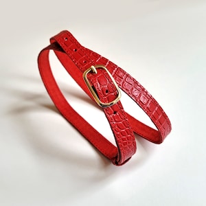 Puede incluir: Un cintur&oacute;n de cuero rojo con una hebilla dorada. El cintur&oacute;n est&aacute; hecho de cuero con estampado de cocodrilo.