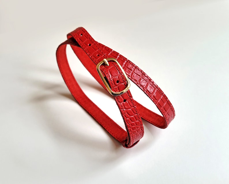 Puede incluir: Un cintur&oacute;n de cuero rojo con una hebilla dorada. El cintur&oacute;n est&aacute; hecho de cuero con estampado de cocodrilo.