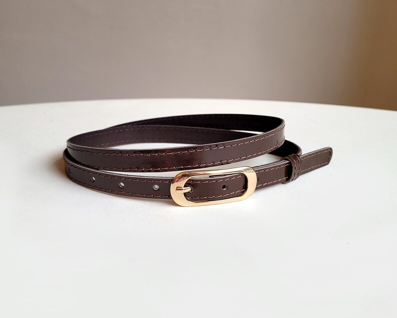 May include: A brown leather belt with a gold-toned buckle. The belt is folded in half and the buckle is visible in the centre of the image.