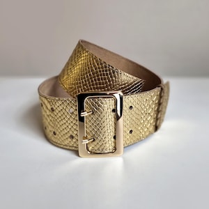 Op de afbeelding: Een brede gouden riem met een gestructureerd slangenleerpatroon en een grote, gepolijste rechthoekige gouden gesp met twee pinnen. De riem heeft twee rijen gaatjes over de lengte en een gladde, lichtgekleurde voering aan de binnenzijde.