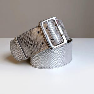 Op de afbeelding: Een brede riem met een metallic zilveren afwerking en een gestructureerd slangenleerpatroon. De riem is voorzien van een grote, rechthoekige, gepolijste zilverkleurige metalen gesp met een doorn, wat zorgt voor een veilige sluiting.