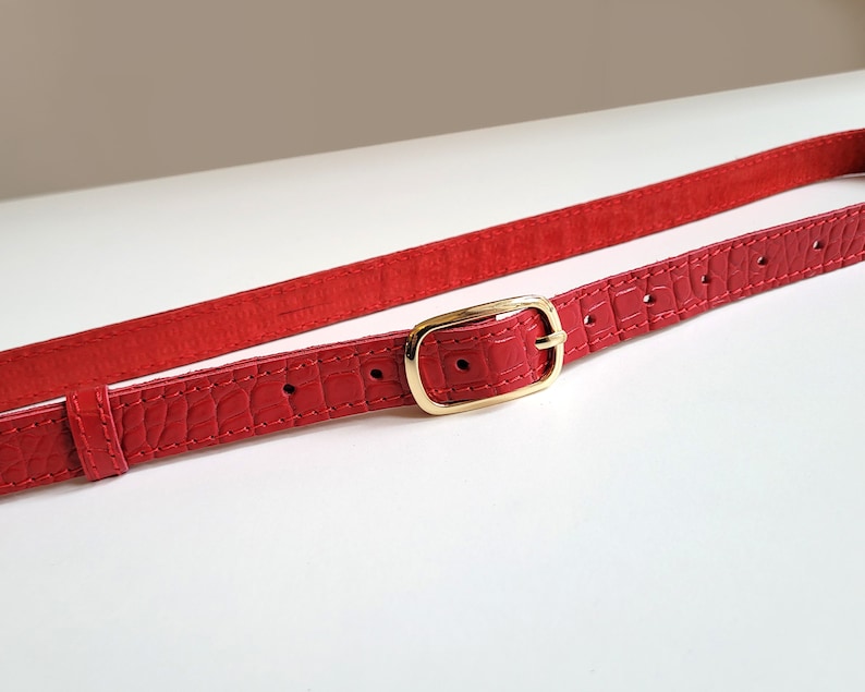 Puede incluir: Un cintur&oacute;n de cuero rojo con una hebilla dorada. El cintur&oacute;n tiene un patr&oacute;n de piel de cocodrilo.