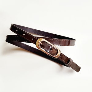 May include: A brown leather belt with a gold-toned oval buckle. The belt is folded in half and the buckle is visible in the centre of the image.