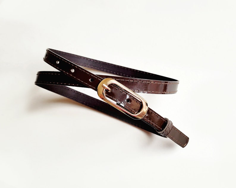 May include: A brown leather belt with a gold-toned oval buckle. The belt is folded in half and the buckle is visible in the centre of the image.