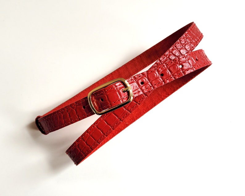 Puede incluir: Un cintur&oacute;n de cuero rojo con una hebilla dorada. El cintur&oacute;n est&aacute; hecho de cuero con estampado de cocodrilo.