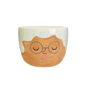 Puede incluir: Un macetero de cerámica con forma de cara de persona con pelo blanco, gafas azules y boca rosa. El macetero es de color marrón claro.