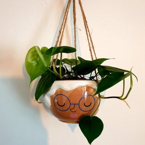 Peut inclure: Une jardinière suspendue avec un pot en céramique blanche en forme de visage de femme aux yeux fermés et à la bouche rose. Le pot est rempli de plantes vertes.