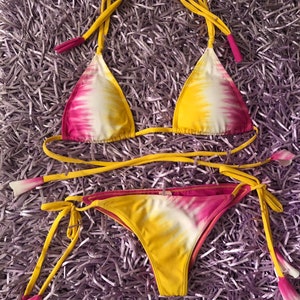 May include: A yellow and pink tie-dye bikini with thin straps and tassels. The bikini top is a triangle style and the bottom is a cheeky style.