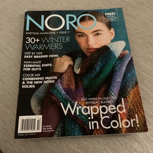 Puede incluir: Portada de la revista "NORO Knitting Magazine Issue 7" con una mujer con un chal de punto colorido. La portada anuncia "30+ Winter Warmers" y "Wrapped in Color!" con información sobre patrones de tejido.