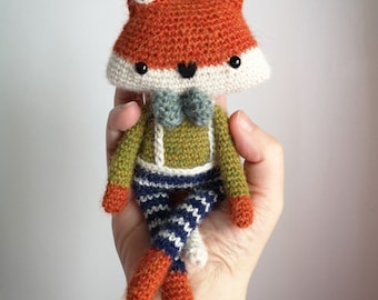 Puede incluir: Un juguete de zorro de crochet que lleva una pajarita verde y azul y pantalones a rayas azules y blancas. El zorro tiene pelo naranja, detalles blancos en la cara y ojos negros.